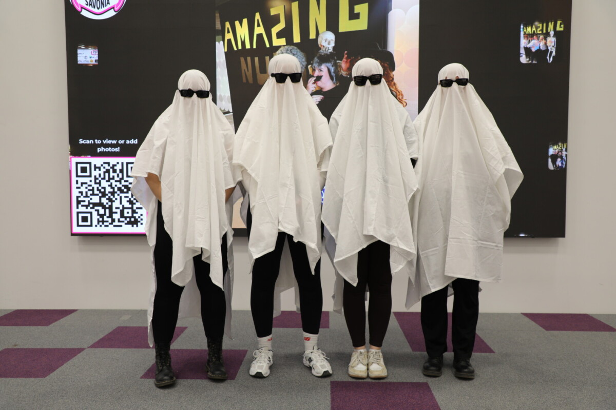 Neljä kummituksiksi pukeutunutta ihmistä seisoo vierekkäin, kukin peitettynä valkoiseen lakanaan ja mustilla aurinkolaseilla. He ovat sisätiloissa violetilla ja harmaalla matolla, ja taustalla on QR-koodi ja värikkäitä julisteita.