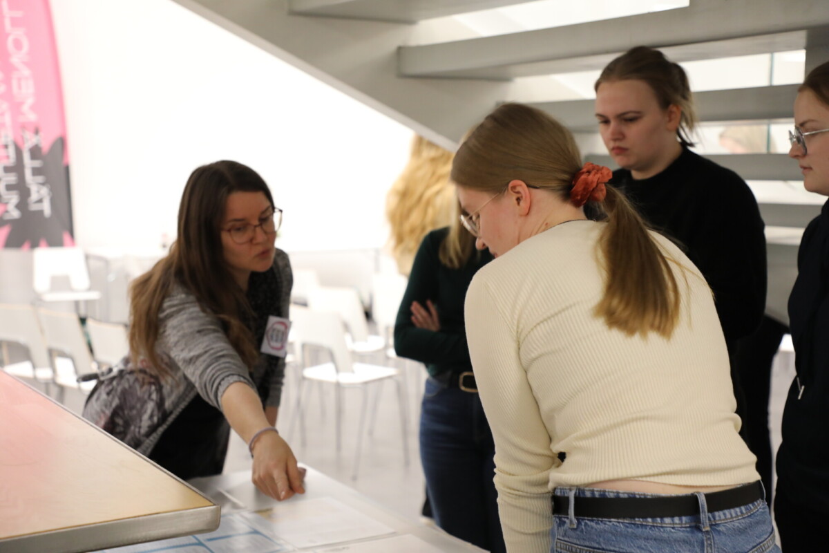 Pitkätukkainen nainen osoittaa pöydällä olevia papereita, kun kolme muuta naista seisoo vieressä ja kuuntelee tarkkaavaisesti valoisassa sisätilassa, jossa on valkoisia tuoleja ja portaikko taustalla.