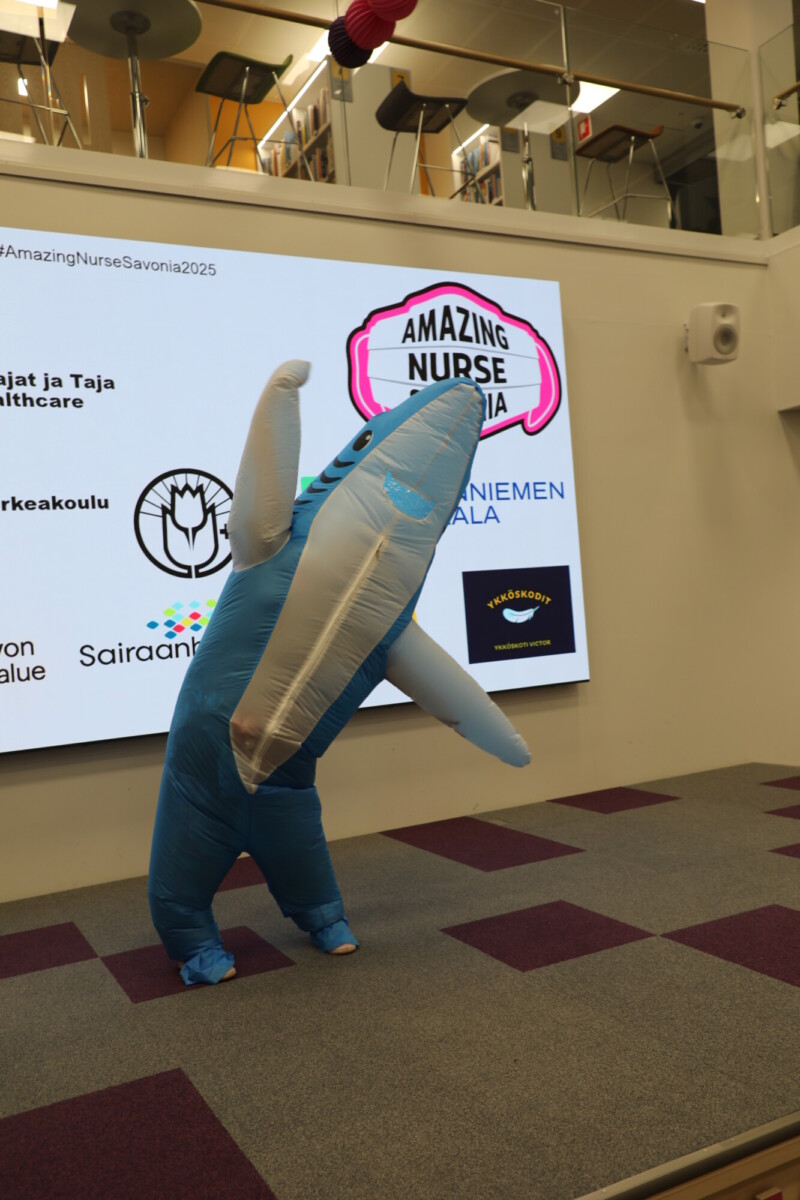 Siniseen puhallettavaan haipukuun pukeutunut henkilö poseeraa toinen evä kohotettuna Amazing Nurse Savonia -logoa ja sponsorilogoja näyttävän suuren näytön edessä modernissa sisätilassa.