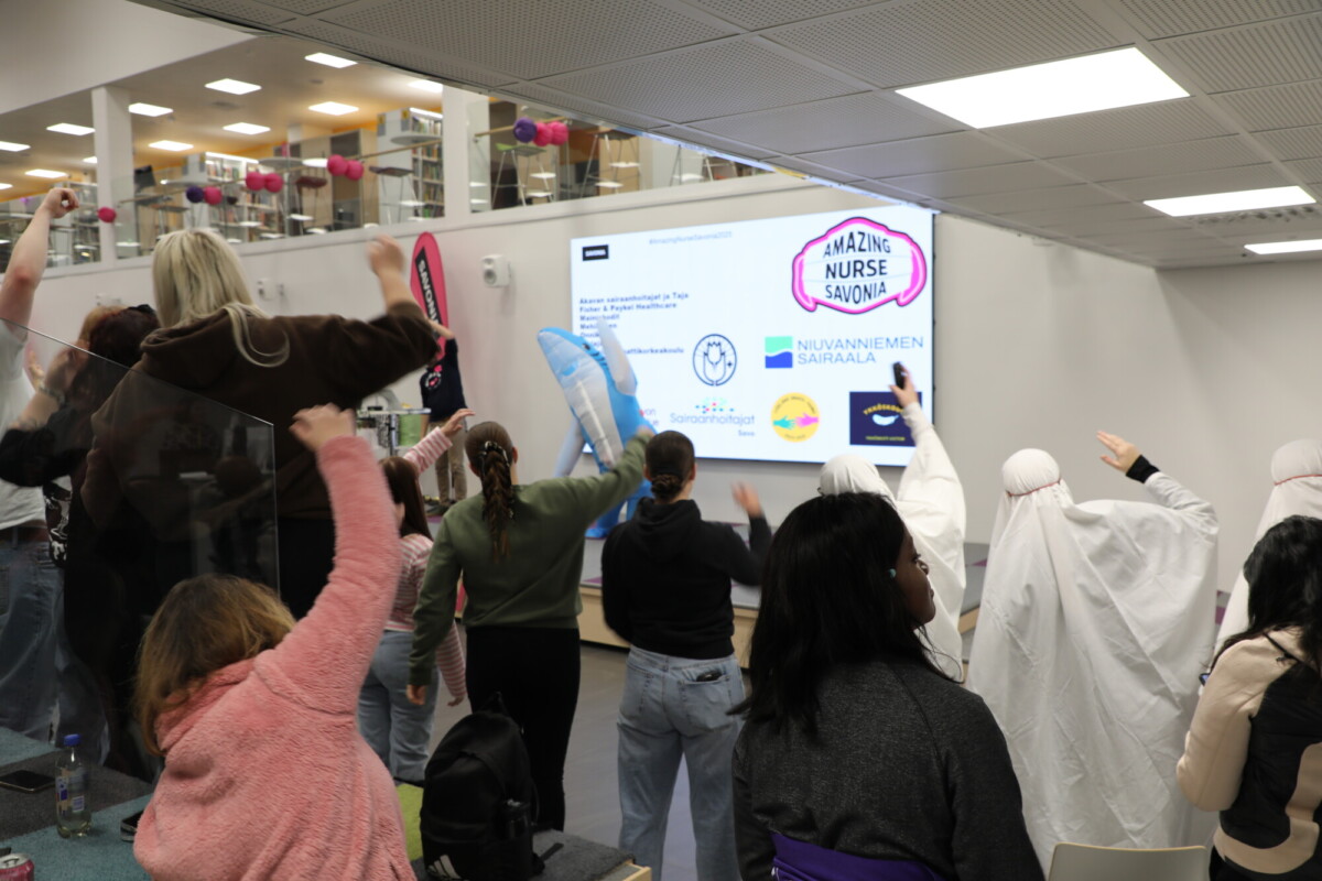 Joukko ihmisiä, joista osa on pukeutunut pukuihin, seisoo sisätiloissa suurta näyttöä vastapäätä, jossa on logoja ja tekstiä, ja nostaa kätensä ylös ikään kuin noudattaisi ohjeita. Huone vaikuttaa kirjastolta tai avoimelta yhteistilalta.