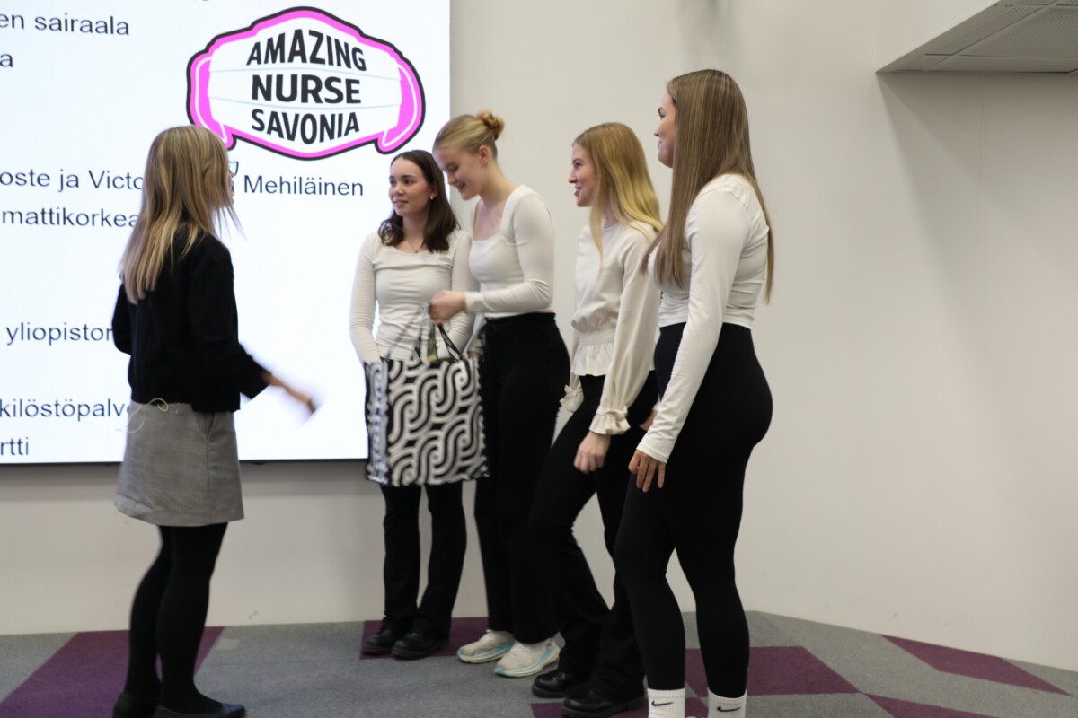 Viisi nuorta naista, joilla kaikilla on valkoiset topit ja tummat alushousut, keskustelevat yhdessä lavalla. Heidän takanaan on valkokangas, jonka puhekuplassa lukee AMAZING NURSE SAVONIA.