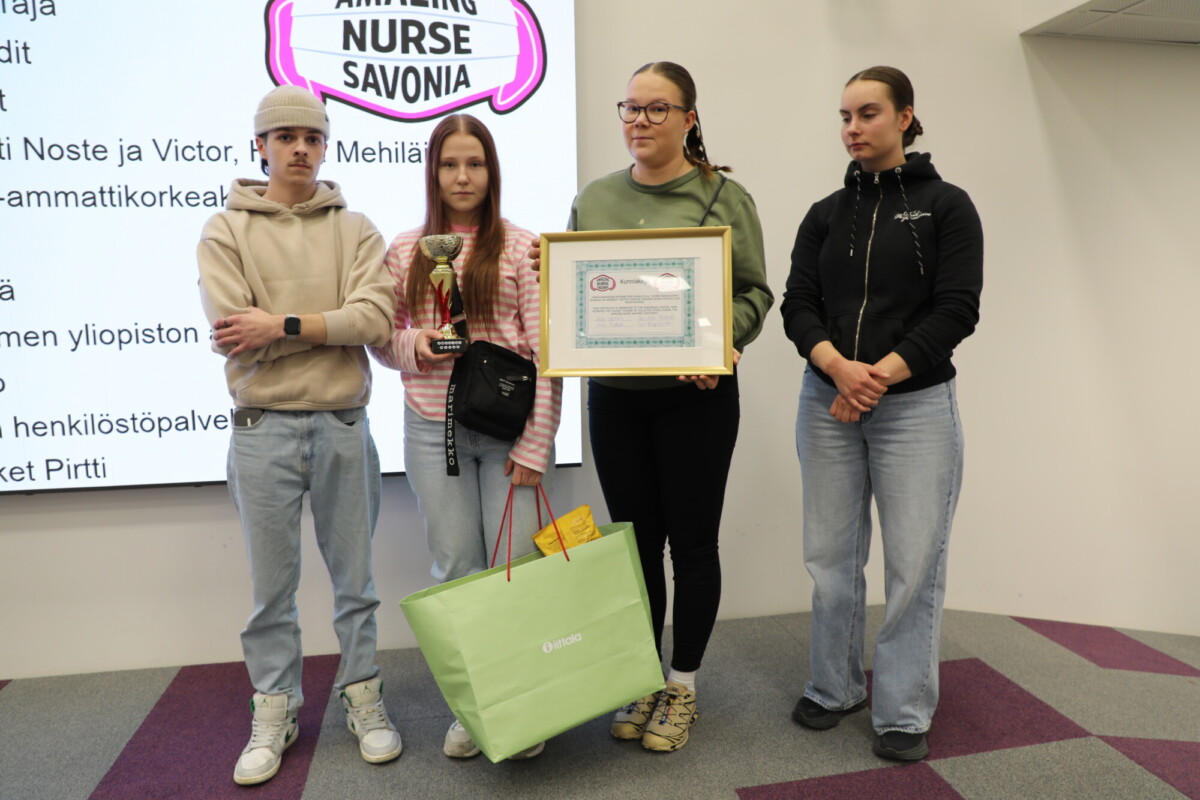 Neljä nuorta seisoo sisätiloissa; kaksi keskellä seisovaa nuorta pitää kädessään pokaalia ja kehystettyä todistusta. Heidän jalkojensa juuressa on suuri vihreä kassi. Heidän takanaan on valkokangas, jossa on teksti AMAZING NURSE SAVONIA.