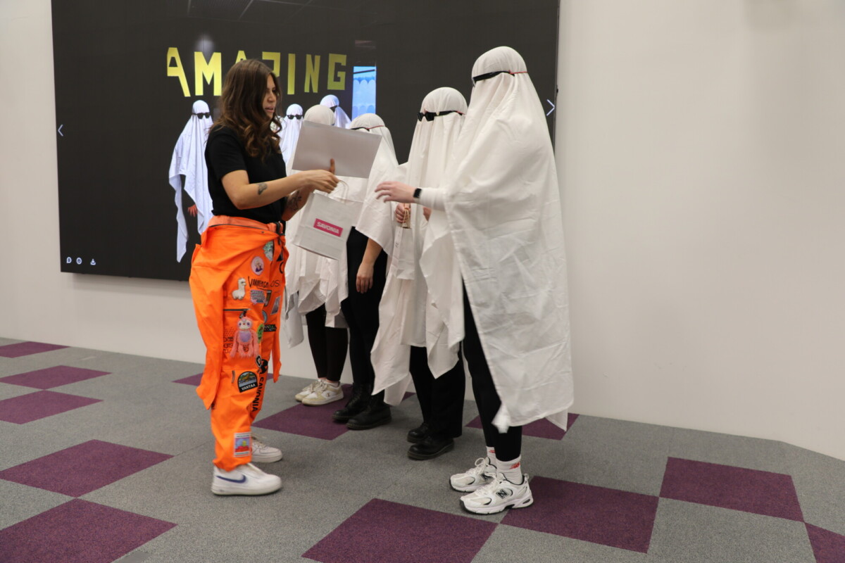 Kirkkaan oransseihin housuihin pukeutunut nainen ojentaa papereita neljälle valkoisiin lakanoihin pukeutuneelle aaveeksi pukeutuneelle ihmiselle, jotka seisovat näytön edessä, jossa lukee AMAZING. Kohtaus tapahtuu sisätiloissa violetilla ja harmaalla matolla.