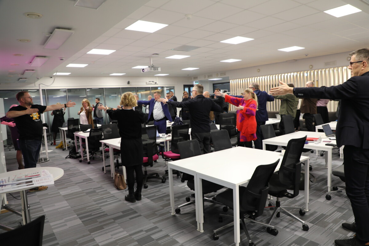 Joukko liikemiesten asuun pukeutuneita ihmisiä seisoo neuvotteluhuoneessa kädet ojennettuina ja osallistuu ryhmäharjoitukseen tai venyttelee pöytä- ja tuolirivien välissä.