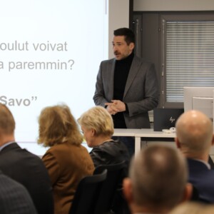 Harmaaseen bleiseriin pukeutunut mies seisoo ja puhuu istuvan yleisön edessä kokoushuoneessa. Hänen takanaan näytöllä näkyy suuri esitysdia, jossa on suomenkielistä tekstiä.