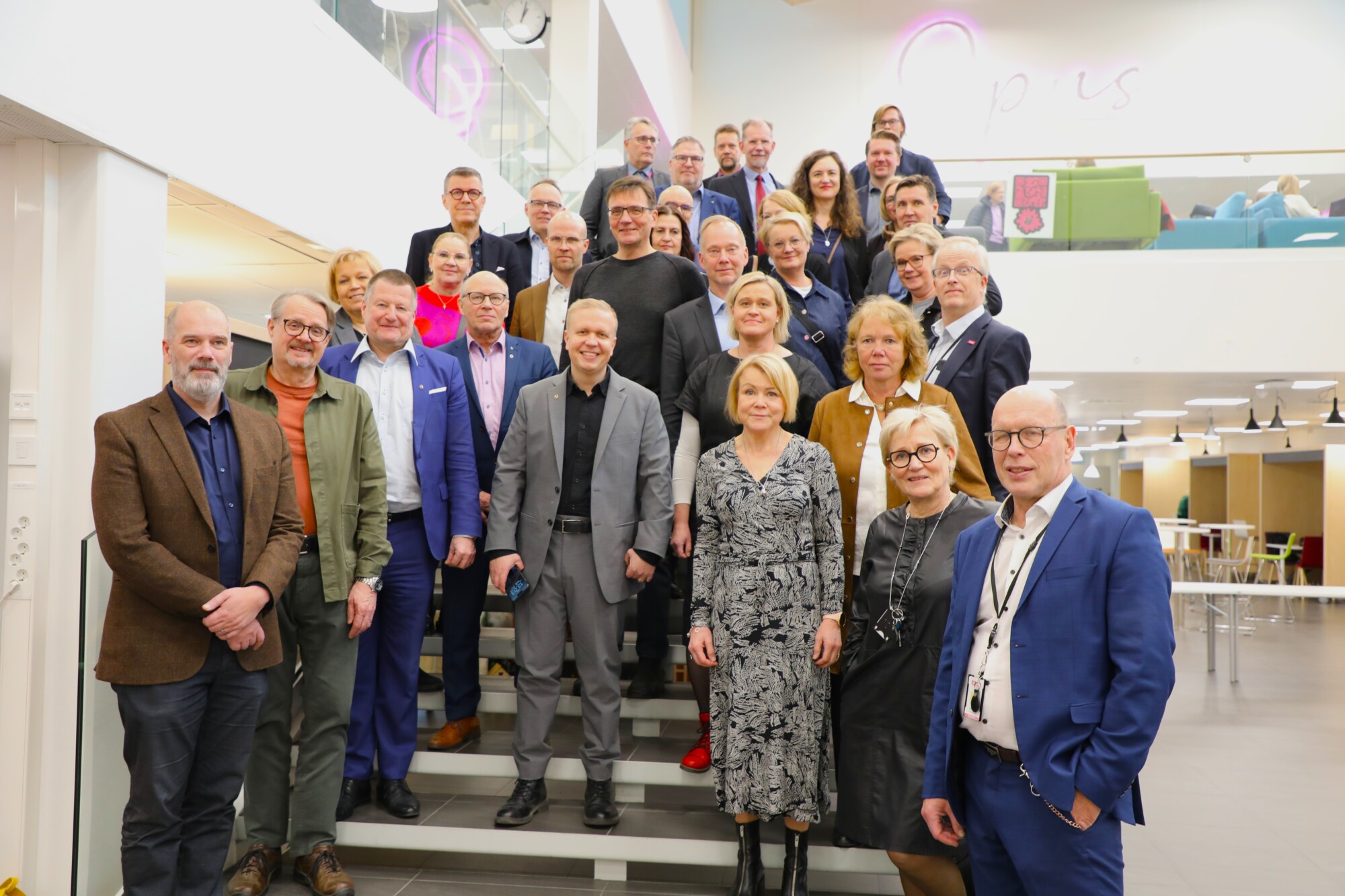 Ryhmä ammattimaisesti pukeutuneita aikuisia poseeraa yhdessä valokuvaa varten portaikossa modernissa, hyvin valaistussa rakennuksessa, jossa on lasia ja avoimia tiloja.