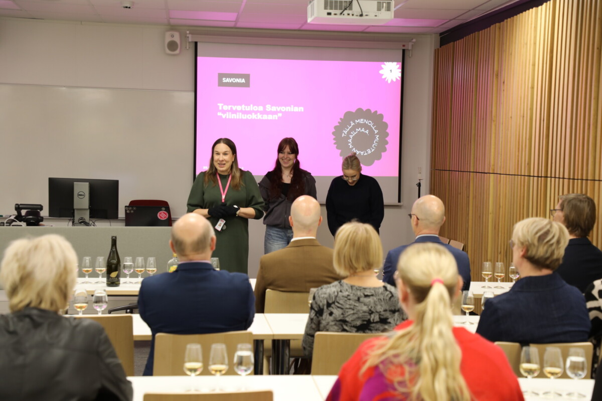 Kolme naista seisoo luokkahuoneen etuosassa esitelmöimässä yleisölle. Heidän takanaan on vaaleanpunainen näyttö, jossa on suomenkielinen teksti. Yleisö istuu, ja heidän edessään olevilla pöydillä on useita viinilaseja.