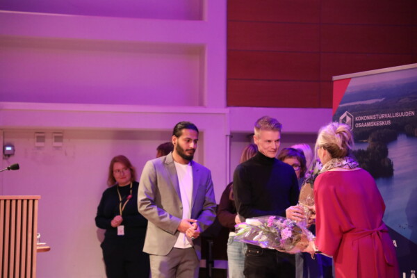 Joukko ihmisiä seisoo sisätiloissa tapahtumassa. Punapukuinen nainen ojentaa kukkakimpun mustaan poolopaitaan pukeutuneelle miehelle, ja muut, muun muassa harmaaseen bleiseriin pukeutunut mies, katselevat ja hymyilevät.