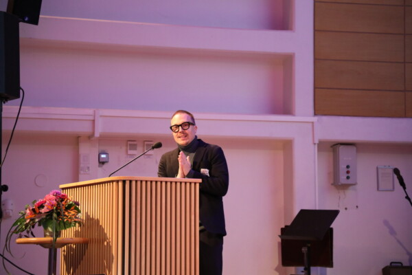 Silmälasit ja tumma puku päällään oleva henkilö seisoo kukkien koristelemalla puisella korokkeella, puhuu mikrofoniin ja hymyilee modernissa sisätilassa, jossa on pehmeä valaistus.