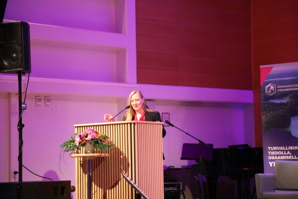 Nainen seisoo puisella korokkeella puhumassa mikrofoniin modernissa sisätiloissa, joissa on violetti valaistus. Hänen vieressään näkyy kukka-asetelma ja pystysuora banneri.