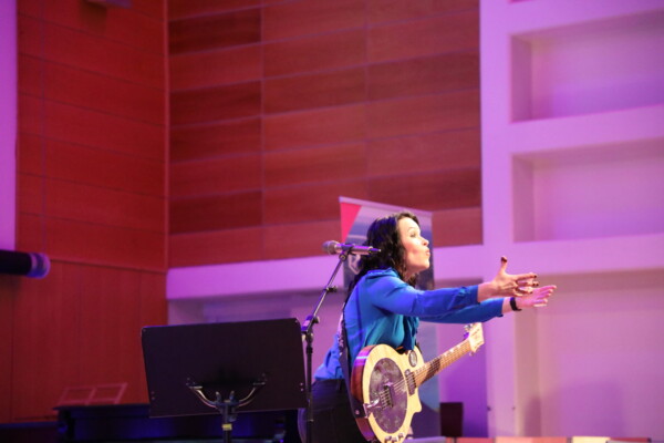 Pitkät tummat hiukset omaava muusikko, jolla on sininen paita, elehtii intohimoisesti avosylin, soittaa banjoa ja laulaa mikrofoniin kirkkaasti valaistulla lavalla, jossa on puisia ja valkoisia paneeleita.