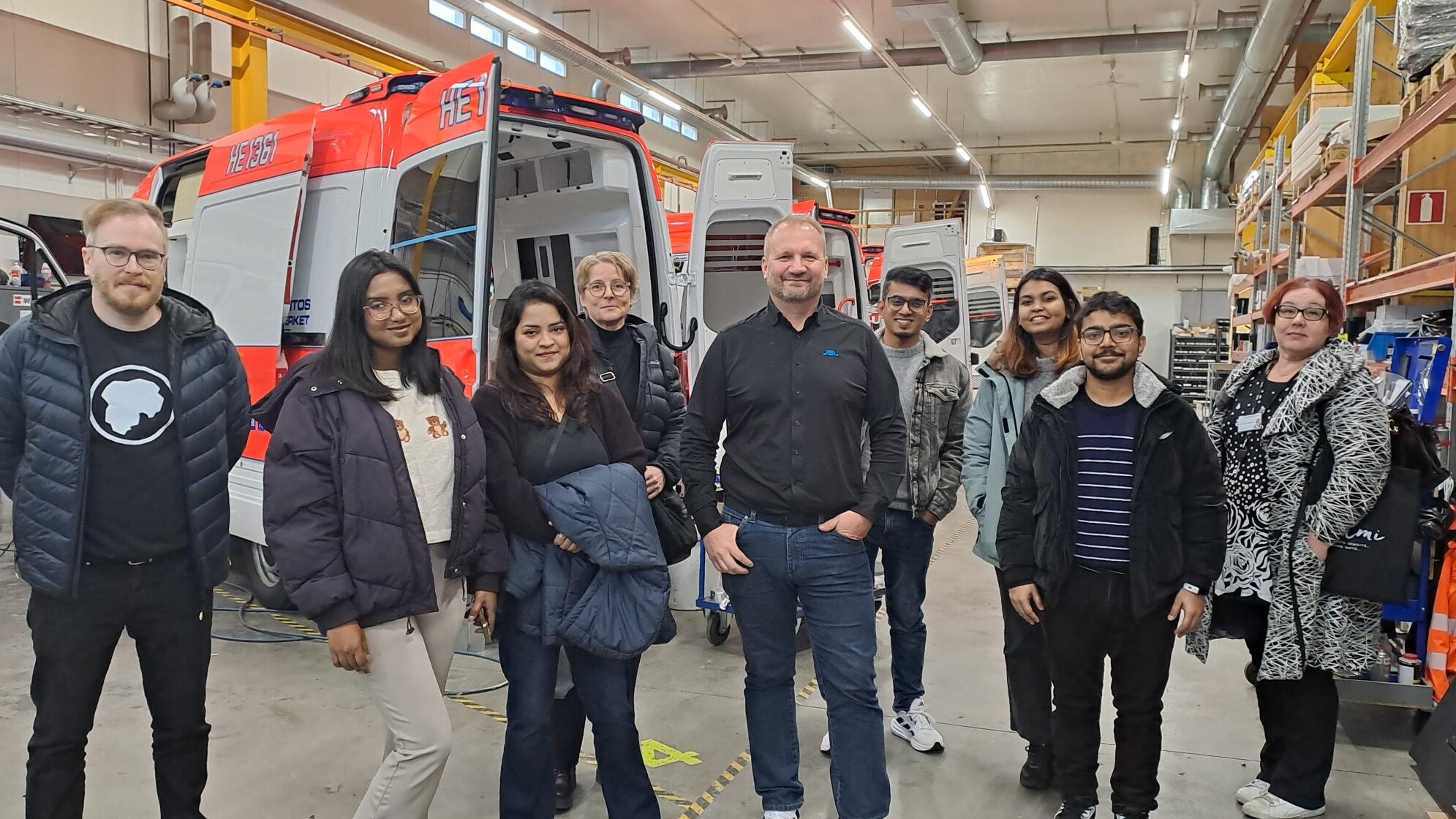 Yhdeksän hengen ryhmä seisoo ja hymyilee suuressa työpajassa, jonka taustalla näkyy osittain koottuja hälytysajoneuvoja ja työkaluja. Ryhmä näyttää poseeraavan valokuvaa varten kierroksen tai vierailun aikana.