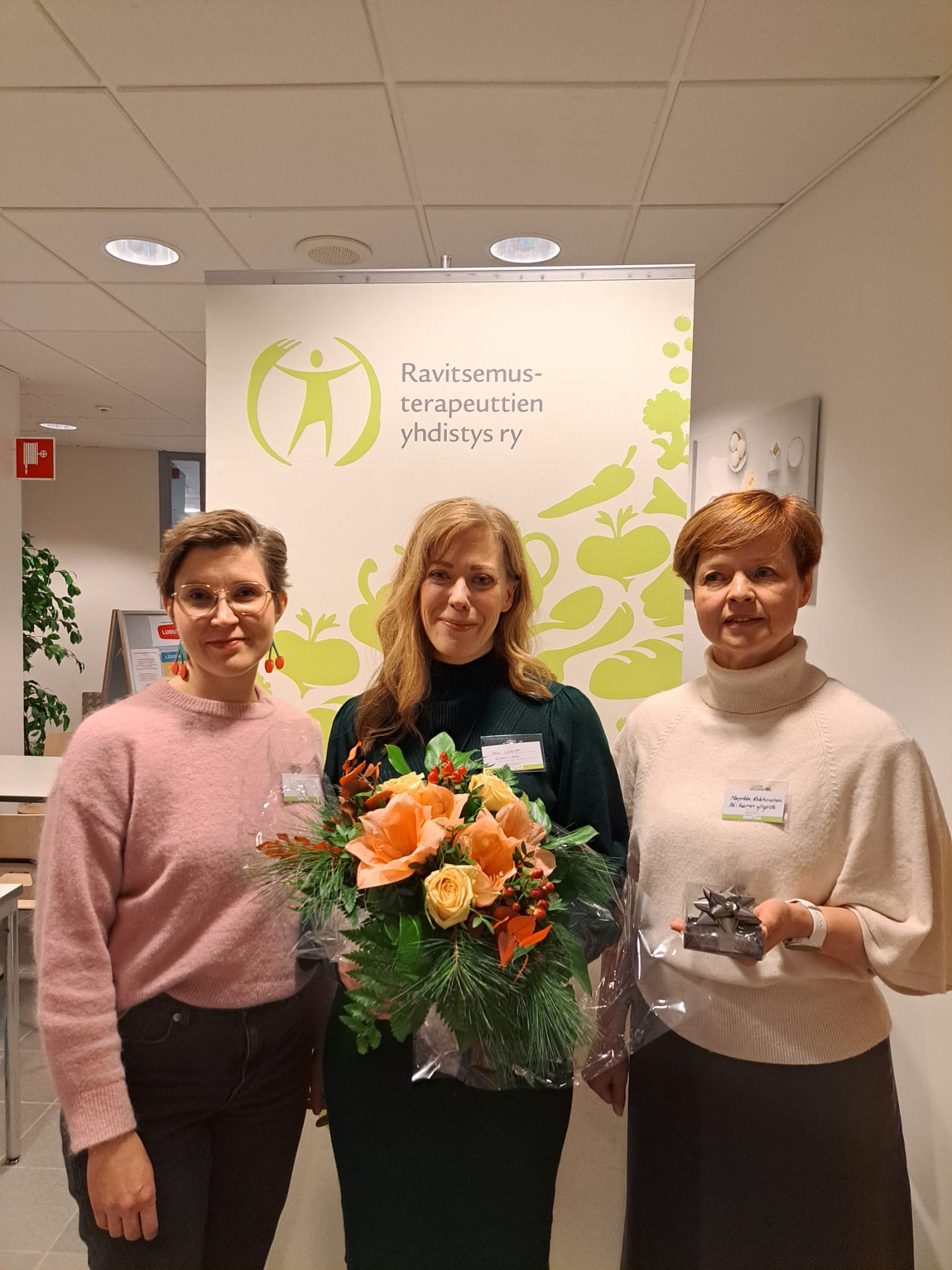 Kolme naista seisoo sisätiloissa suomenkielisen kyltin edessä. Keskellä olevalla naisella on kädessään oranssi kukkakimppu, ja naiset molemmin puolin hymyilevät kameralle.