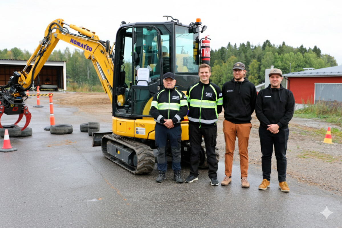 Neljä miestä seisoo pienen keltaisen Komatsu-kaivinkoneen edessä märällä päällystetyllä pinnalla, ja taustalla on liikennekartioita, renkaita ja puita; kaikilla on työvaatteet yllään ja he hymyilevät kameralle.