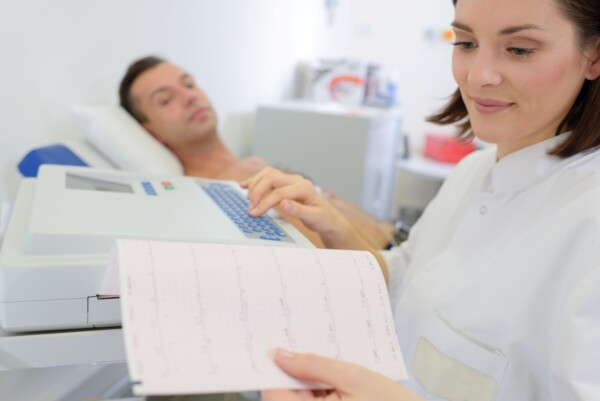 Valkoiseen takkiin pukeutunut terveydenhuollon ammattilainen tarkastelee EKG-tulostetta sairaalasängyllä makaavan potilaan vieressä, joka on kytketty EKG-laitteeseen.