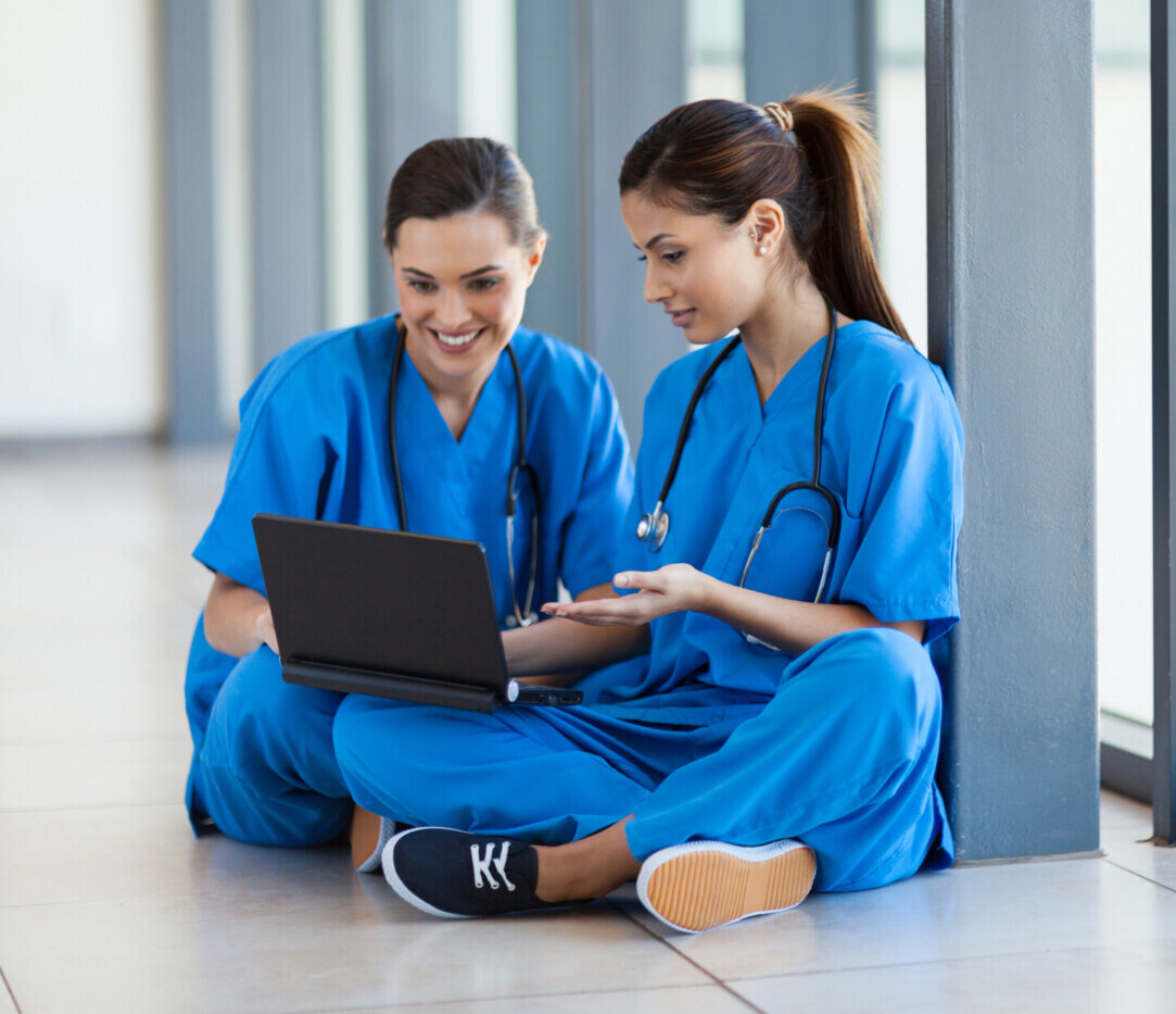 Kaksi sinisiin hoitohaalareihin pukeutunutta naista istuu lattialla suurten ikkunoiden äärellä, katselee kannettavaa tietokonetta ja hymyilee. Molemmilla on stetoskoopit kaulassaan, ja he näyttävät keskustelevan jostain näytöllä näkyvästä asiasta.