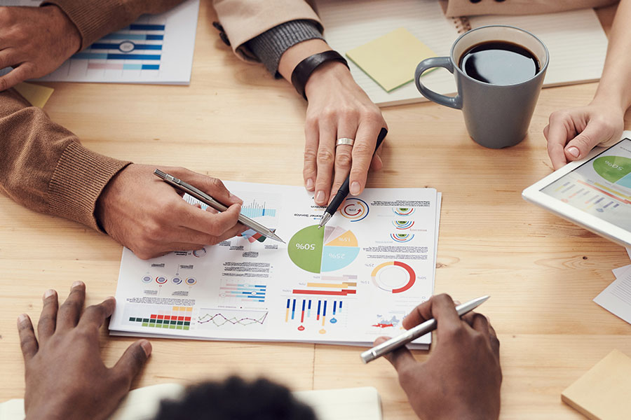 Neljä ihmistä istuu pöydän ympärillä tarkastelemassa värikkäitä kaavioita ja kaavioita paperilla ja tabletilla. Kädet osoittavat asiakirjoja kynillä, ja lähellä on kuppi kahvia. Pöytä on puinen ja siinä on hajallaan toimistotarvikkeita.