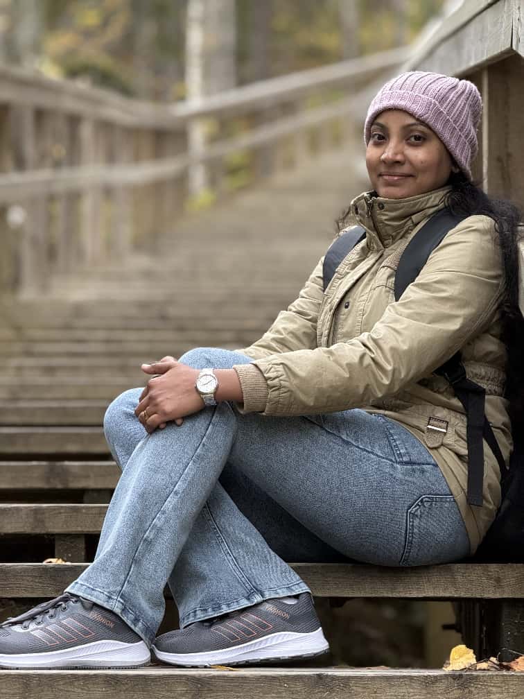 Nainen, jolla on vaaleanpunainen pipo, khaki-takki, siniset farkut ja lenkkarit, istuu ulkona puuportailla ja hymyilee hieman kameralle, ja taustalla näkyy puita hämärtyneenä.