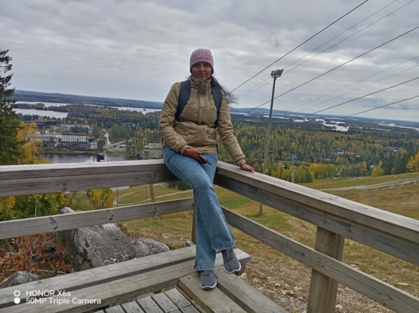 Vaaleanpunaiseen pipoon ja takkiin pukeutunut nainen istuu puukaiteella, josta avautuu näkymä maisemaan, jossa on syksyisiä puita, vettä ja rakennuksia pilvisen taivaan alla. Hän hymyilee ja hänellä on reppu yllään.