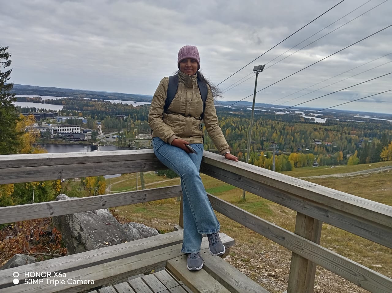 Vaaleanpunaiseen pipoon ja takkiin pukeutunut nainen istuu puukaiteella, josta avautuu näkymä maisemaan, jossa on syksyisiä puita, vettä ja rakennuksia pilvisen taivaan alla. Hän hymyilee ja hänellä on reppu yllään.