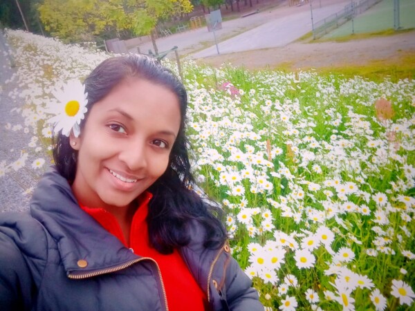 Nainen, jolla on päivänkakkara hiuksissaan, hymyilee kameralle ja seisoo kukkivien valkoisten päivänkakkaroiden kentän edessä. Hänellä on tumma takki ja punainen toppi, ja taustalla näkyy vihreitä puita ja tenniskenttä.