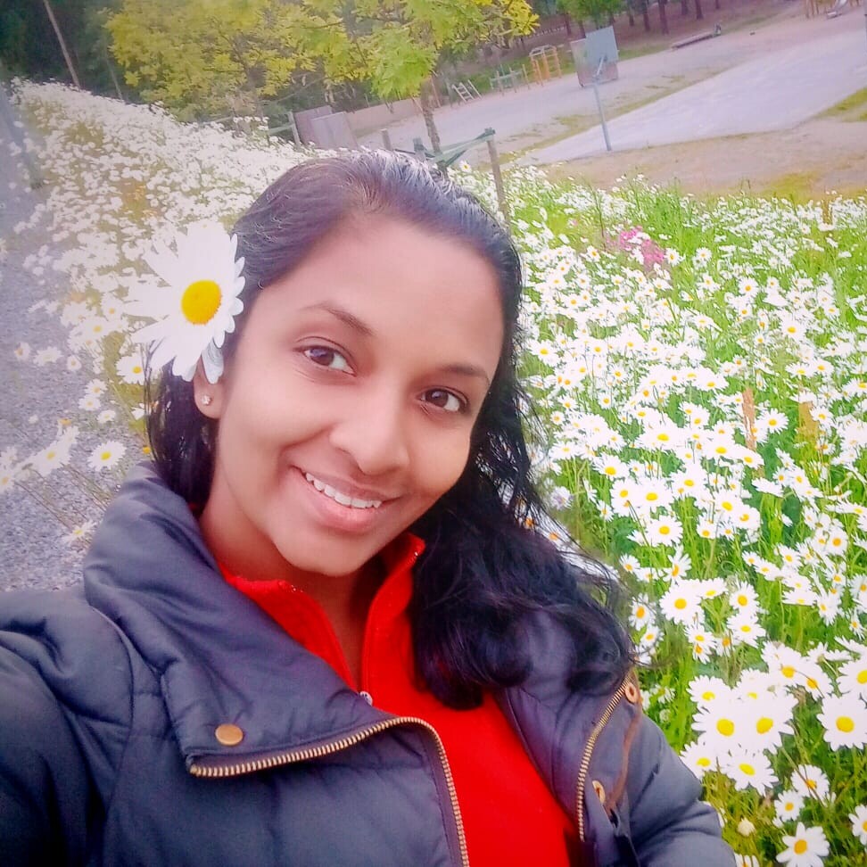 Nainen, jolla on päivänkakkara hiuksissaan, hymyilee kameralle ja seisoo kukkivien valkoisten päivänkakkaroiden kentän edessä. Hänellä on tumma takki ja punainen toppi, ja taustalla näkyy vihreitä puita ja tenniskenttä.