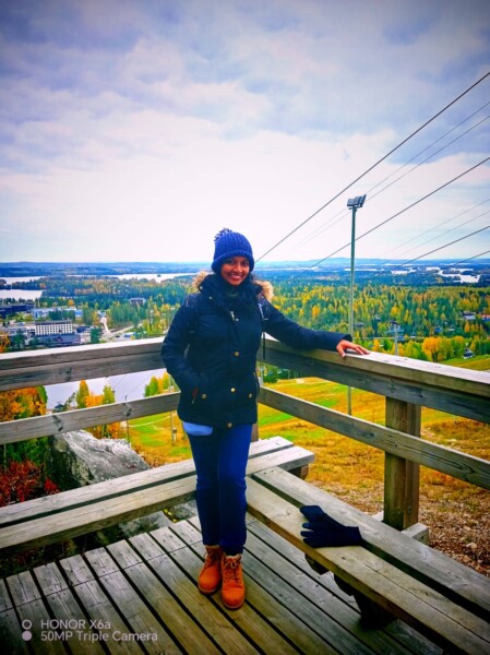 Tummaan talvitakkiin, siniseen hattuun ja saappaisiin pukeutunut henkilö seisoo hymyillen puisella korokkeella, josta avautuu näkymä maisemaan, jossa on syksyisiä puita, rakennuksia ja pilvinen taivas.