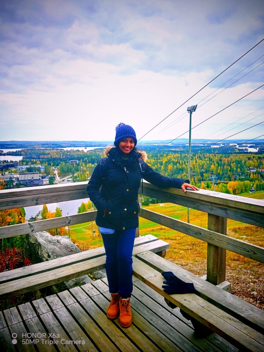 Tummaan talvitakkiin, siniseen hattuun ja saappaisiin pukeutunut henkilö seisoo hymyillen puisella korokkeella, josta avautuu näkymä maisemaan, jossa on syksyisiä puita, rakennuksia ja pilvinen taivas.