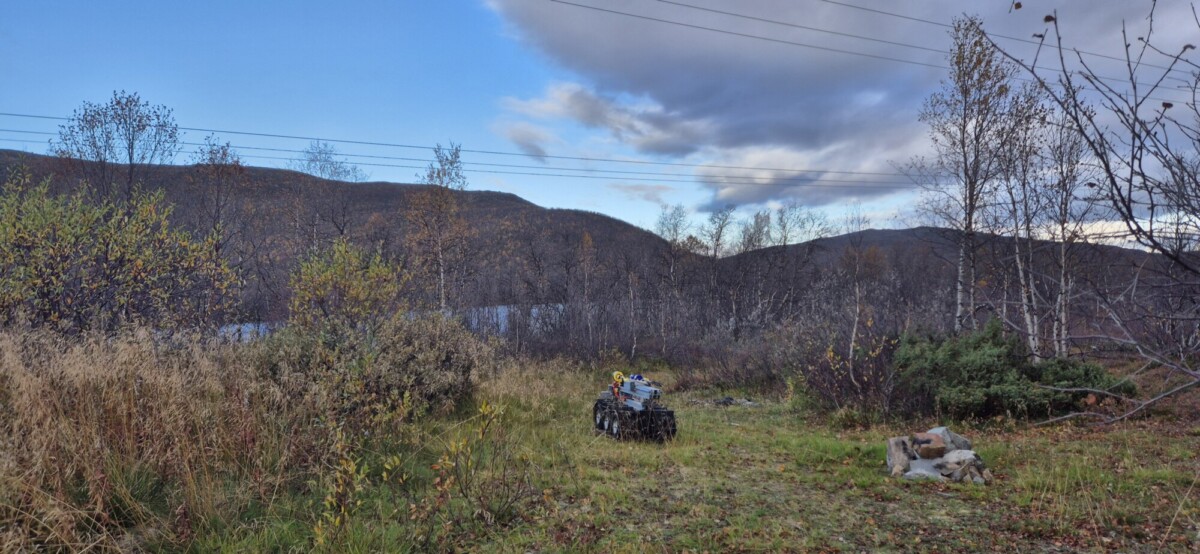 Pieni robottiajoneuvo istuu ruohikkoisella polulla syksyisten puiden ja pensaiden ympäröimänä, ja taustalla on kukkuloita ja pilvinen sininen taivas. Kuvan yläosan poikki kulkee sähkölinjoja.