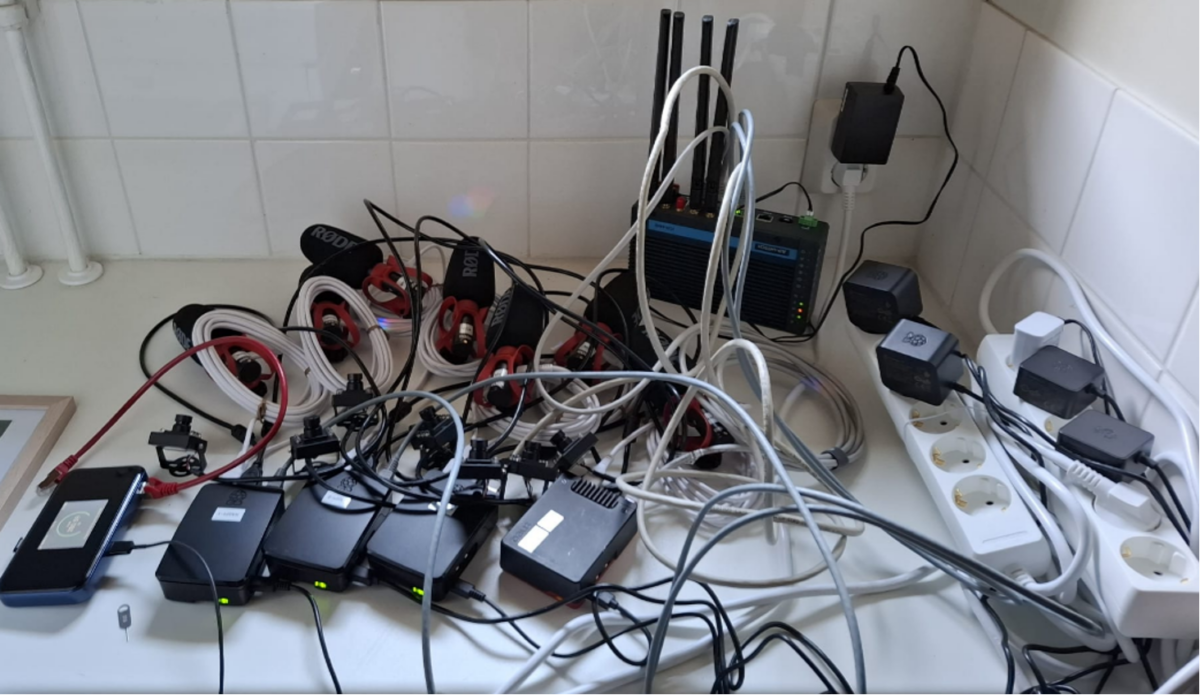 Sotkuinen työtaso, jossa on useita pistorasioita, sekaisin olevia kaapeleita, useita elektronisia laitteita, USB-keskittimiä, reitittimiä antenneineen ja pistorasioihin kytkettyjä verkkosovittimia. Alue näyttää olevan täynnä teknisiä laitteita.