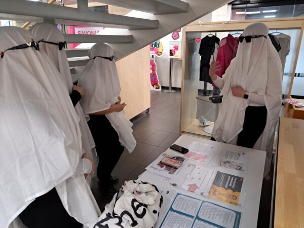 Kolme kummituksiksi pukeutunutta ihmistä, joilla on valkoiset lakanat ja aurinkolasit, seisovat paperipöydän vieressä, kun taas toinen samanlaiseen asuun pukeutunut henkilö elehtii pöydän vieressä, kaikki sisätiloissa portaikon alla.