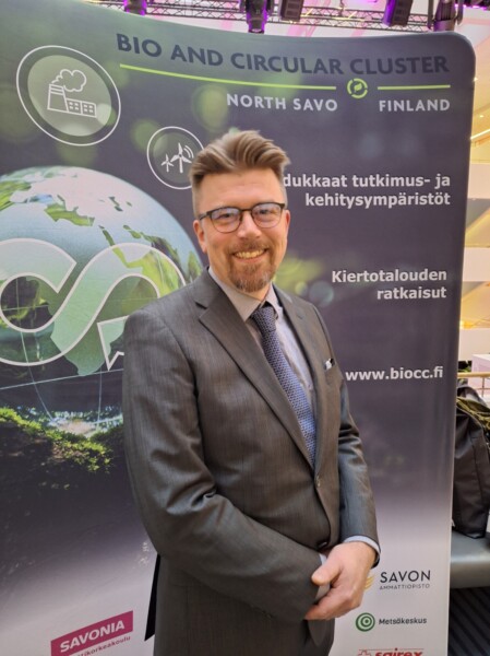 Hymyilevä pukuun ja silmälaseihin pukeutunut mies seisoo bio- ja kiertotalousklusterin näytön edessä, jossa on suomenkielisiä tekstejä ja logoja, maapallografiikka ja tietoa bio- ja kiertotalousratkaisuista.