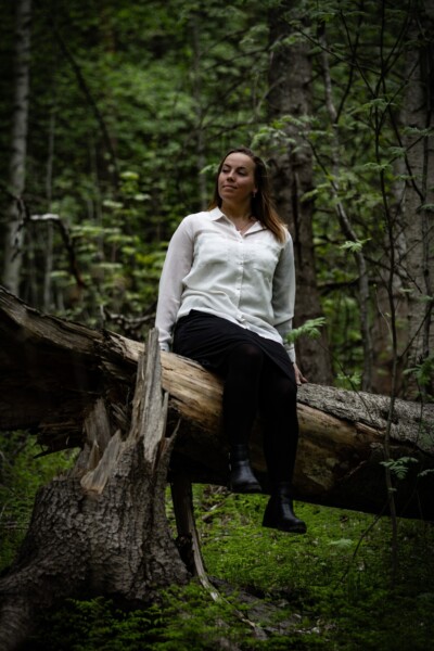 Valkoiseen puseroon ja mustaan hameeseen pukeutunut nainen istuu kaatuneen puunrungon päällä vehreässä metsässä tyytyväisen ja mietteliään näköisenä.