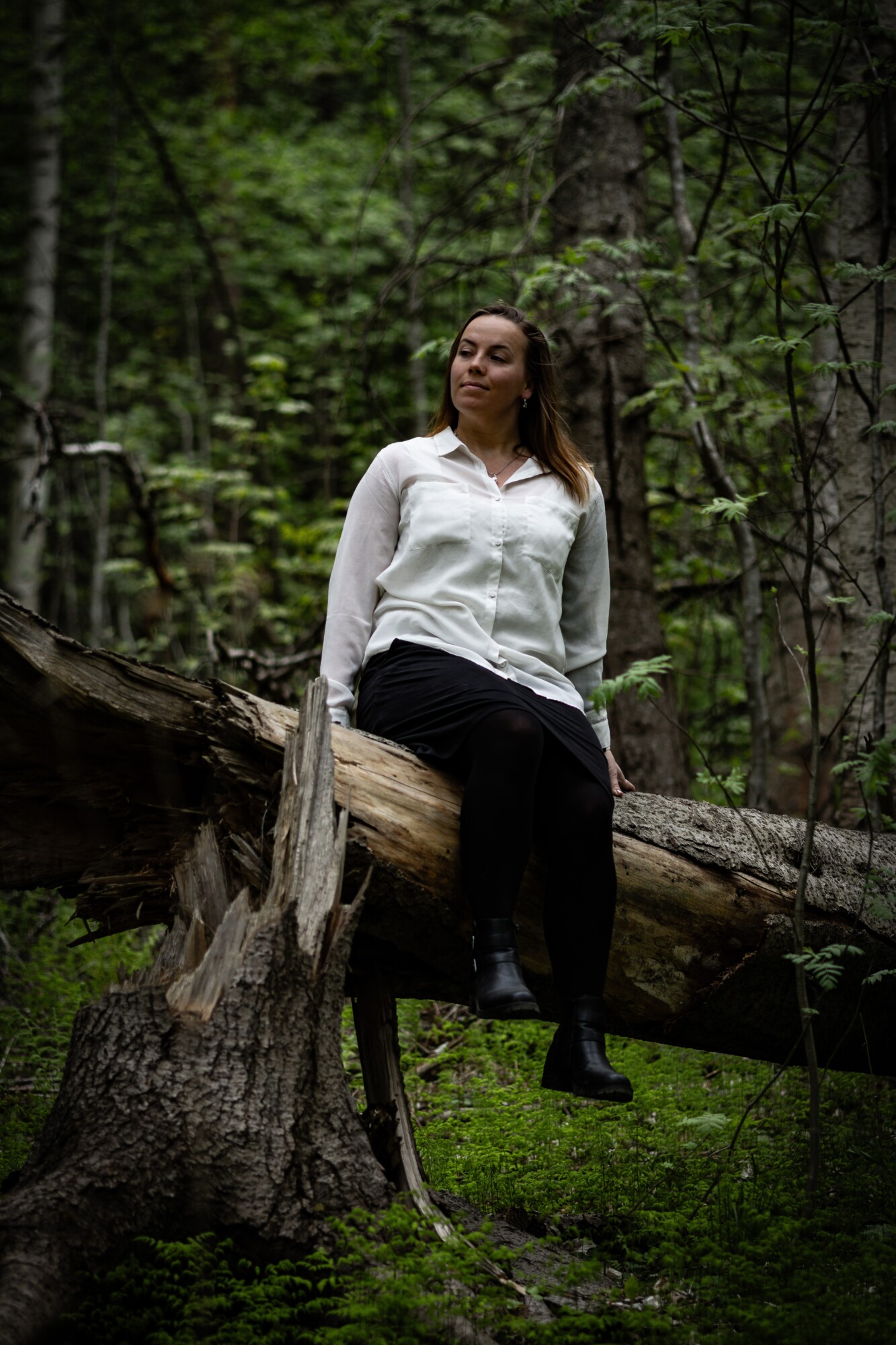 Valkoiseen puseroon ja mustaan hameeseen pukeutunut nainen istuu kaatuneen puunrungon päällä vehreässä metsässä tyytyväisen ja mietteliään näköisenä.