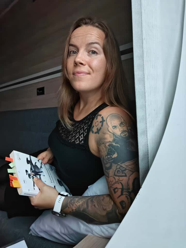 Pitkätukkainen, tatuoitu nainen istuu ikkunan ääressä hymyillen hieman ja pitelee kädessään kirjaa, jossa on värikkäitä muistilappuja. Hänellä on hihaton musta toppi ja valkoinen kello.