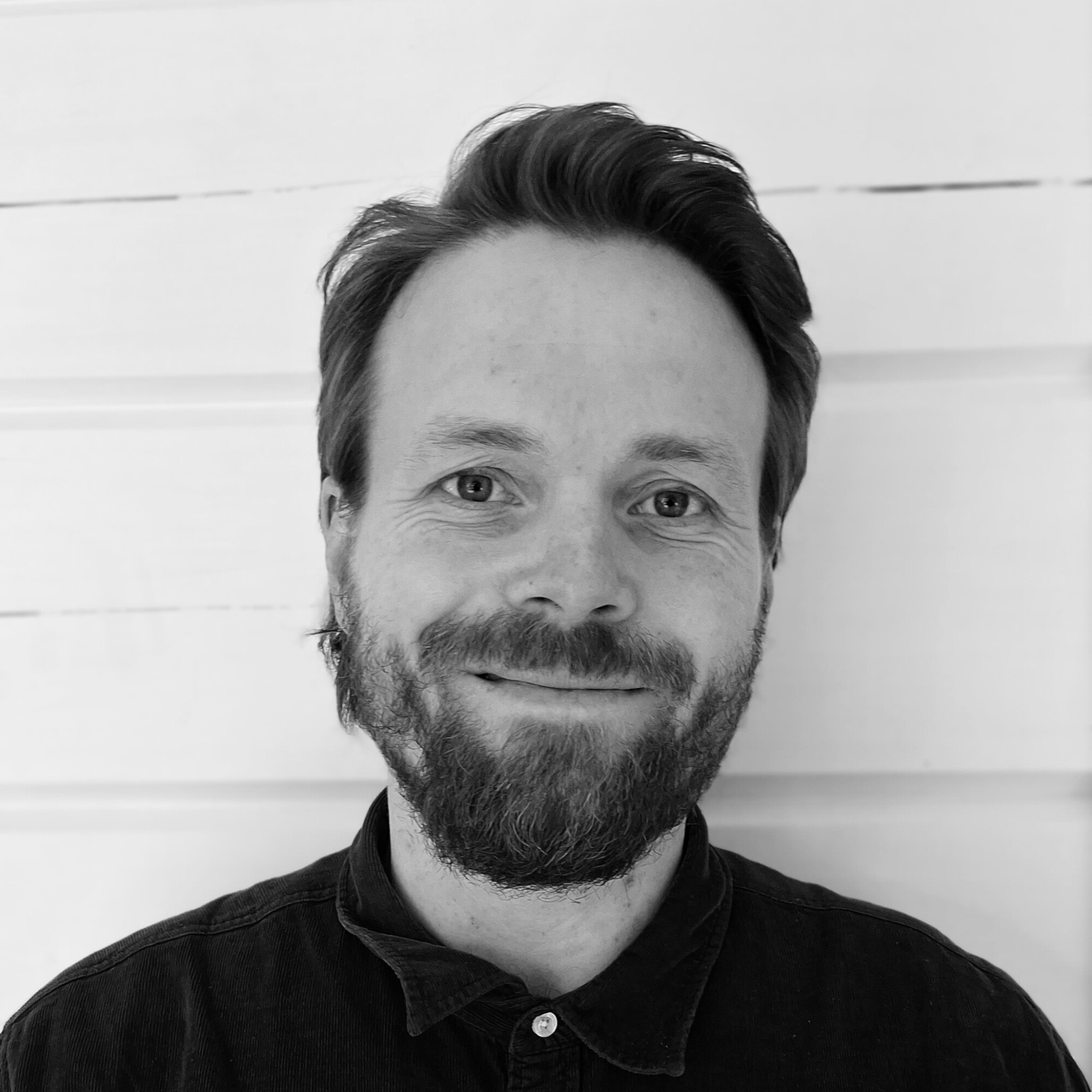 Partainen ja viiksekäs mies, jolla on tumma nappipaita, hymyilee kameralle valkoisen puuseinän edessä.