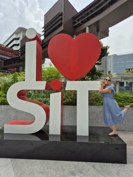 Siniseen mekkoon pukeutunut nainen poseeraa leikkisästi suuren I ♥ SIT -veistoksen vieressä ulkona, taustalla on moderneja rakennuksia ja vehreyttä.