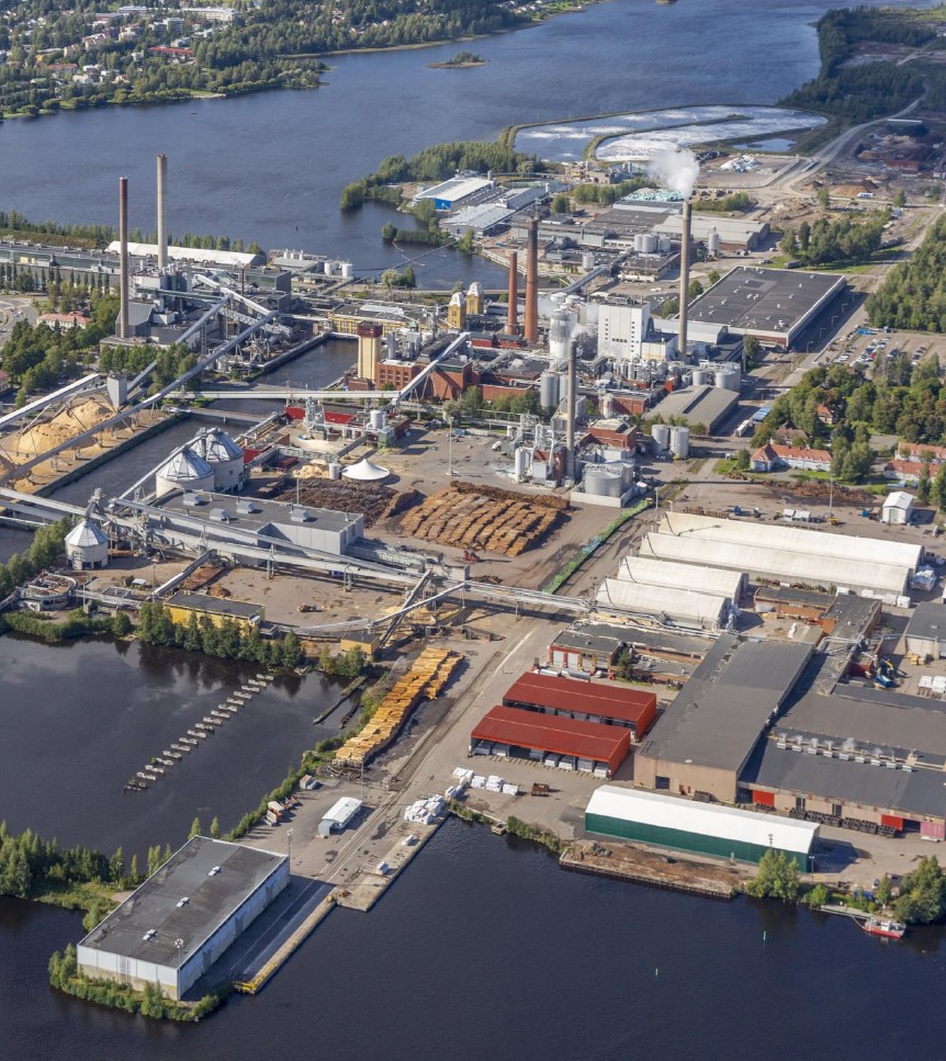 Ilmakuva vesistön rannalla sijaitsevasta teollisuuskompleksista, jossa on tehtaita, savupiippuja, varastoja ja raaka-ainepinoja, joita ympäröivät puut ja läheinen asuinalue.