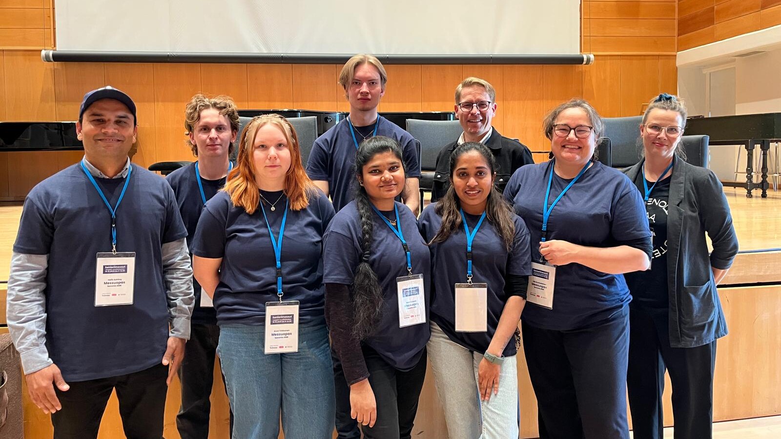 Yhdeksän hengen ryhmä seisoo sisätiloissa puisella lavalla, kaikilla on laivastonsiniset paidat ja nimilaput, ja he hymyilevät kameralle. Taustalla näkyy suuri valkokangas ja puinen panelointi.