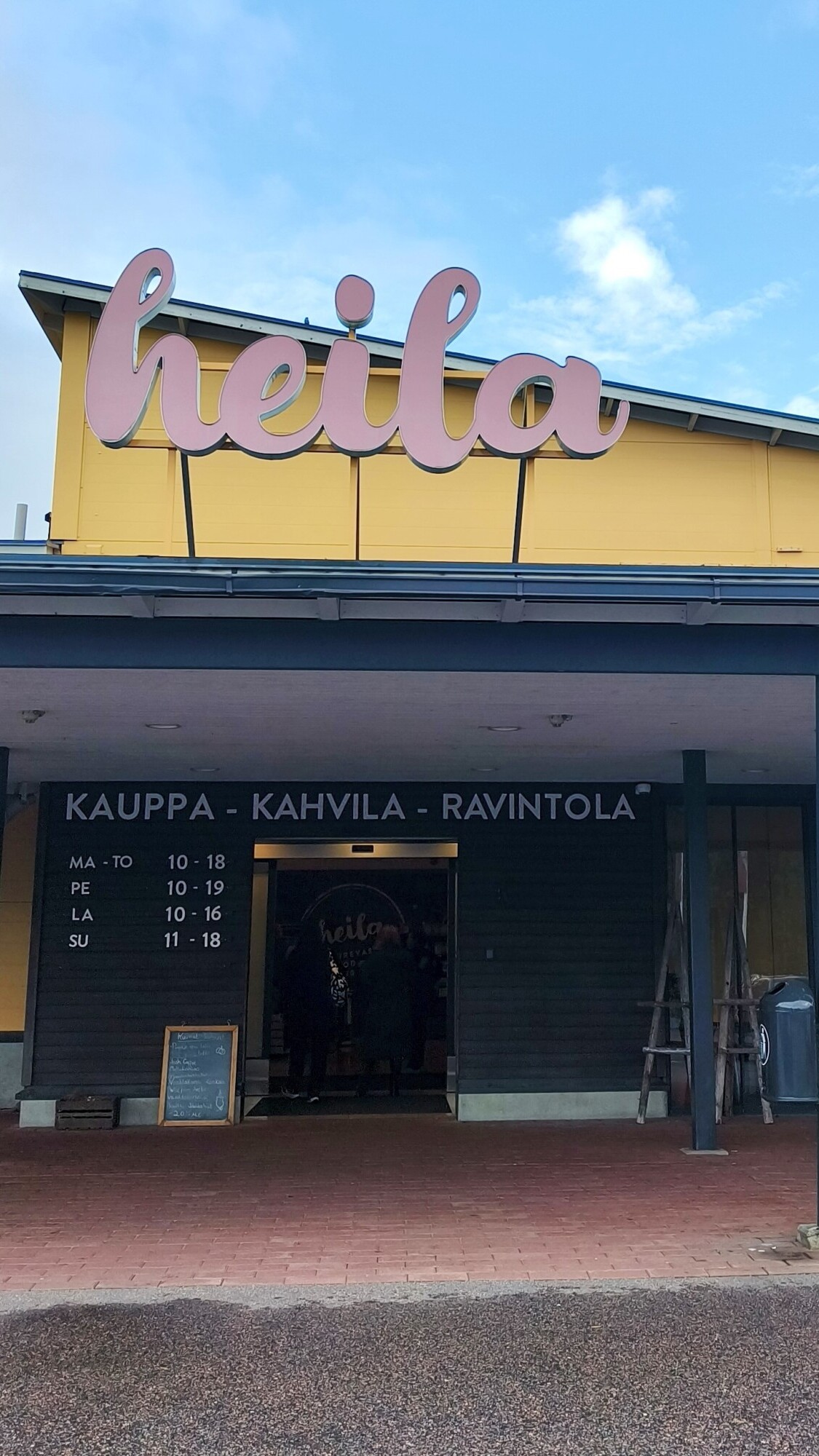 Rakennus, jonka sisäänkäynnin yläpuolella on suuri vaaleanpunainen kyltti, jossa lukee heila. Sen alapuolella suomenkieliset sanat mainostavat kauppaa, kahvilaa ja ravintolaa, ja aukioloajat on lueteltu mustassa seinätaulussa. Taivas on osittain pilvinen.