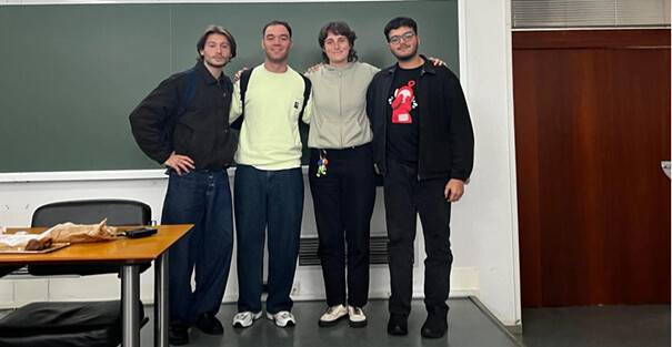 Neljä ihmistä seisoo vierekkäin luokkahuoneessa ja hymyilee kameralle. Heidän edessään on pulpetti, jossa on papereita, ja heidän takanaan on vihreä liitutaulu. Heillä on kädet toistensa ympärillä.