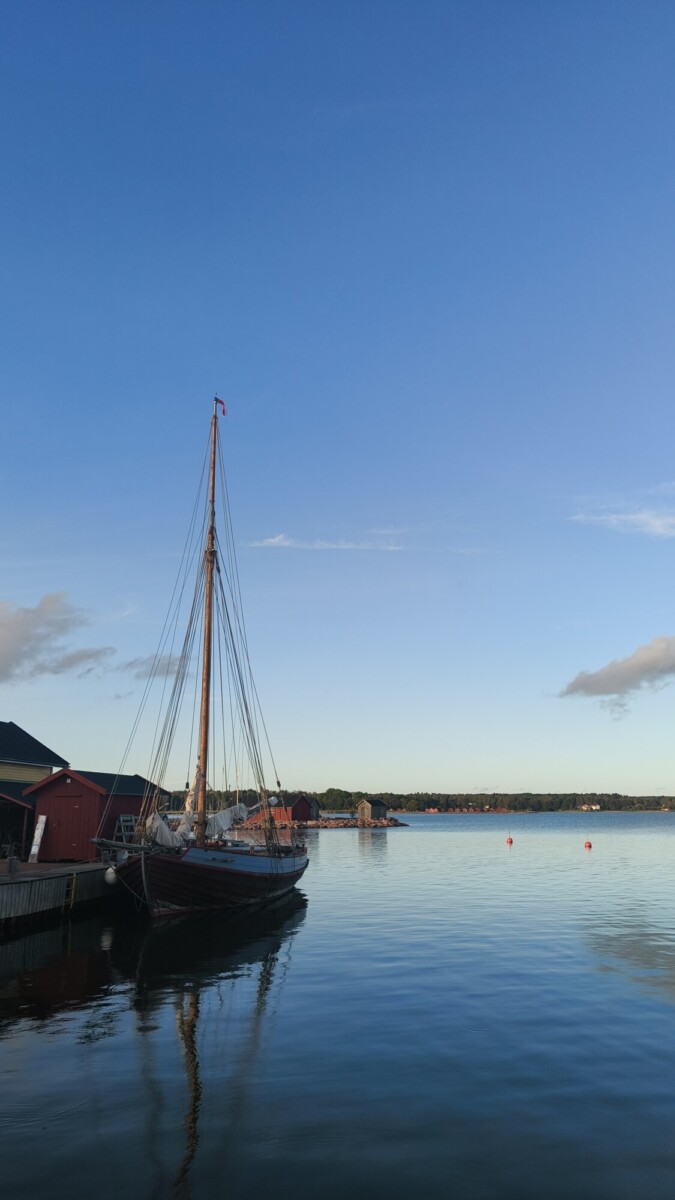 Purjevene, jossa on korkea masto, on telakoituna punaisen puurakennuksen vieressä tyynellä vedellä kirkkaalla sinisellä taivaalla, ja horisontissa näkyy kaukana rantaviiva ja muutama pilvi.