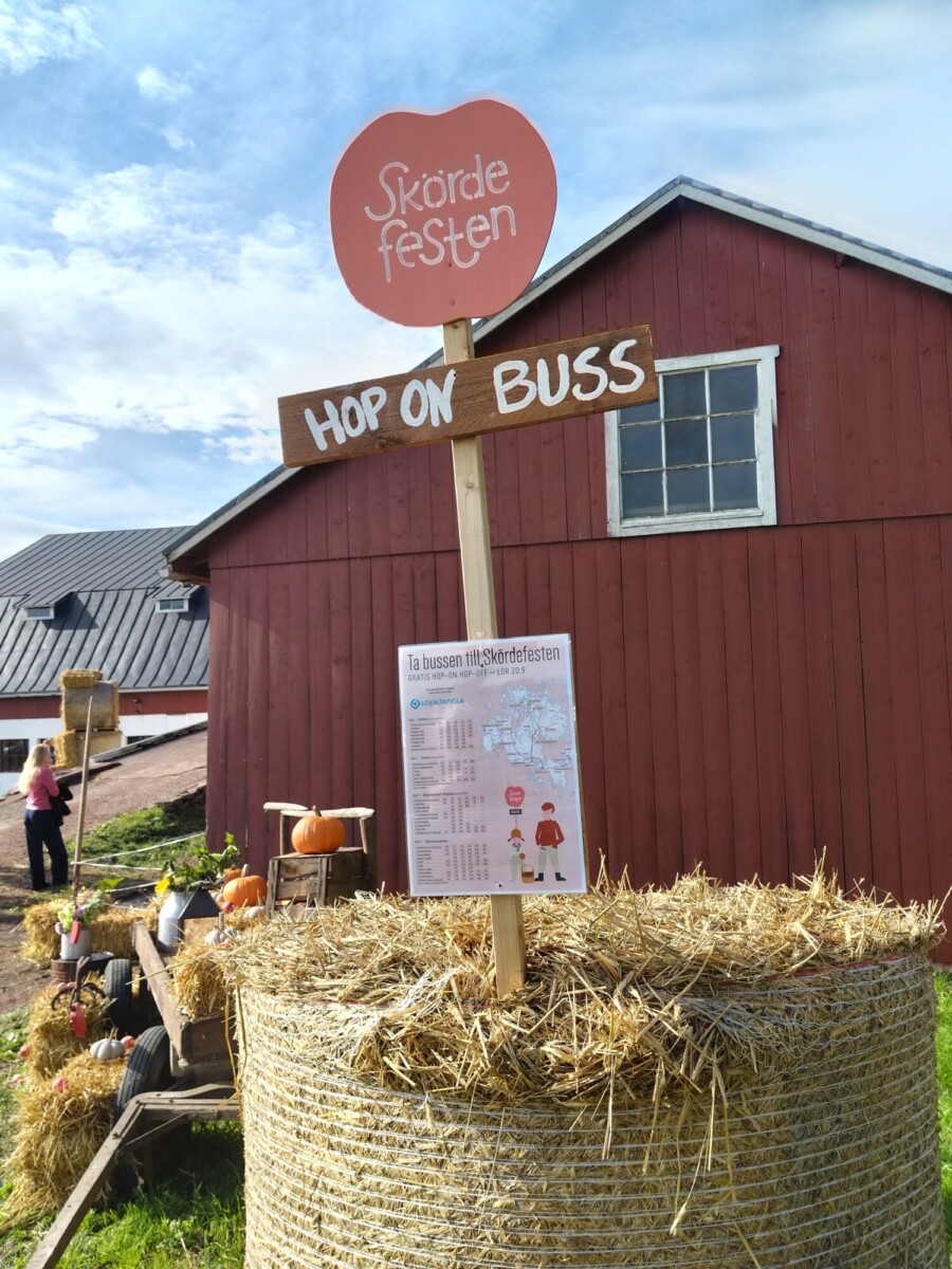 Punaisen ladon edessä seisoo heinäpaali, jossa on puinen kyltti, jossa lukee Hop On Buss ja Skördefesten. Kurpitsat ja festivaalijuliste koristavat maisemaa ja luovat juhlavan syystunnelman.