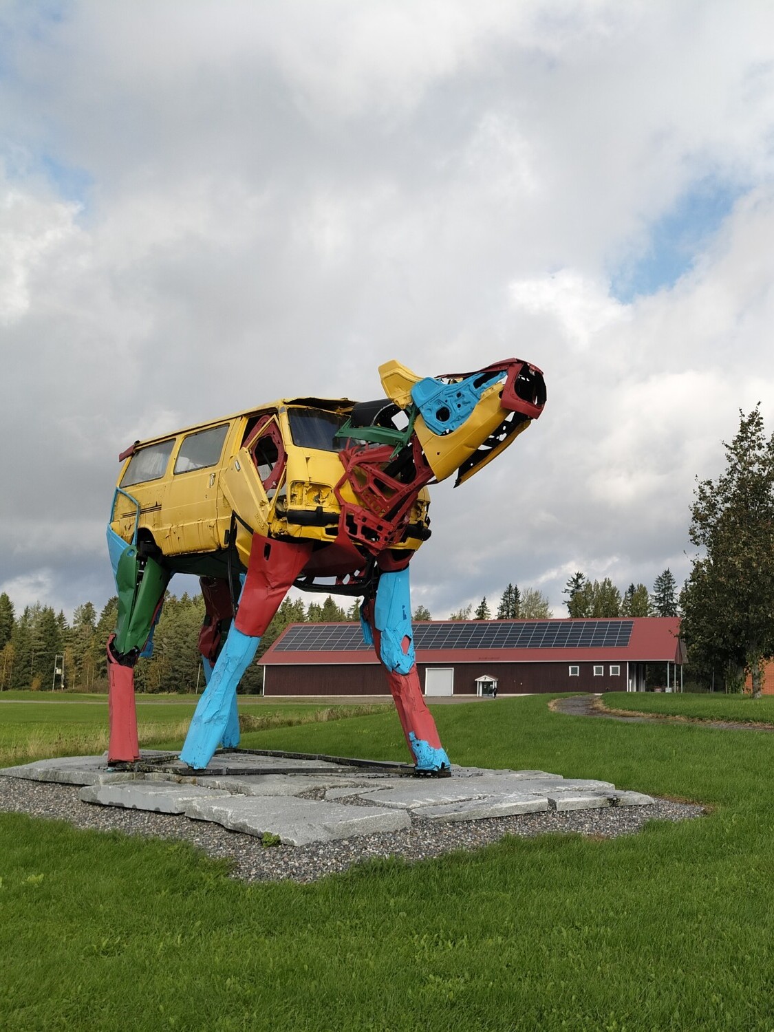 Auton osista tehty värikäs koiran veistos seisoo betonialustalla nurmikentällä, ja taustalla on punainen tiilirakennus ja puita osittain pilvisen taivaan alla.