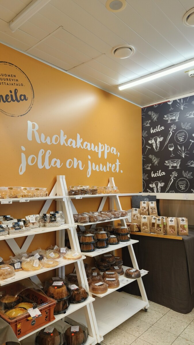 Leipomomyymälän sisustus, jossa on hyllyjä, joissa on erilaisia leipiä ja leivonnaisia. Oranssilla seinällä on suomenkielinen teksti Ruokakauppa, jolla on juuret, ja oikealla on taulutyylinen seinä kuvituksineen.