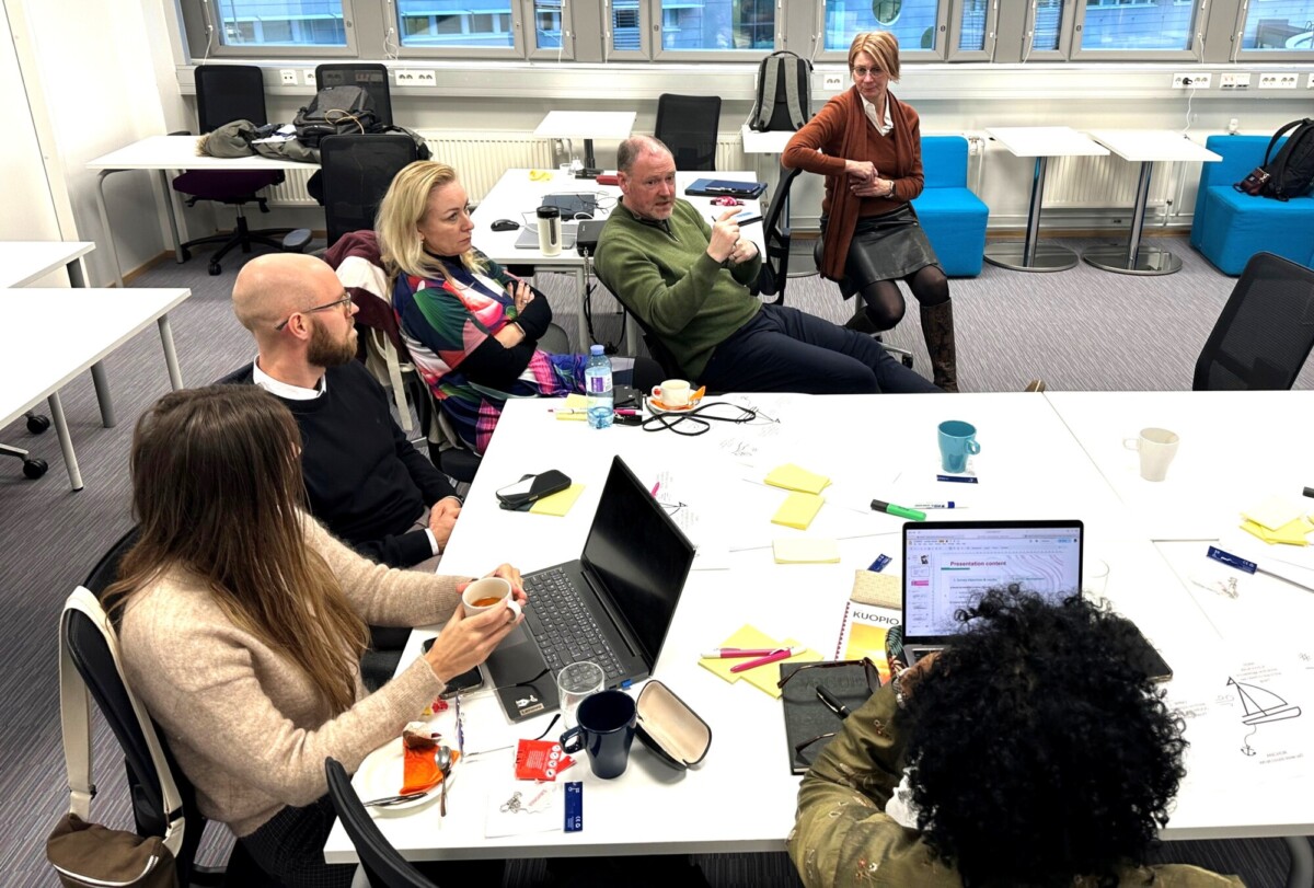 Kuusi ihmistä istuu ja seisoo valkoisen pöydän ympärillä valoisassa toimistossa keskustellen. Pöydällä on hajallaan kannettavia tietokoneita, kahvikuppeja, papereita, kyniä ja muistilappuja.