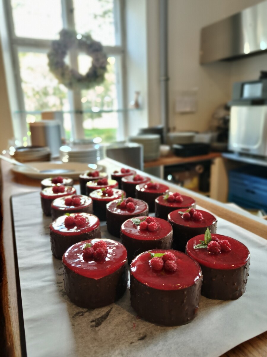 Rivit pyöreitä suklaakakkuja, joiden päällä on kiiltävä punainen vadelmakuorrutus ja tuoreita vadelmia, on aseteltu pergamenttipaperille valoisassa keittiössä, jonka taustalla on ikkuna.