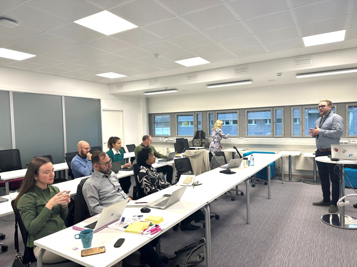 Mies seisoo ja puhuu modernin, valoisan luokkahuoneen edessä, kun taas useat aikuiset istuvat pöydissä kannettavien tietokoneiden, vihkojen ja kahvin kanssa ja kuuntelevat tarkkaavaisesti. Suuret ikkunat reunustavat yhtä seinää.