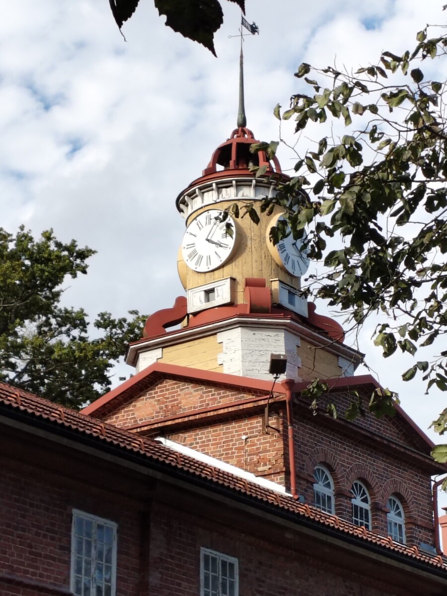 Lähikuva punatiilisestä rakennuksesta, jonka kellotornissa on neljä suurta valkoista kellotaulua, puiden ja osittain pilvisen taivaan ympäröimänä.