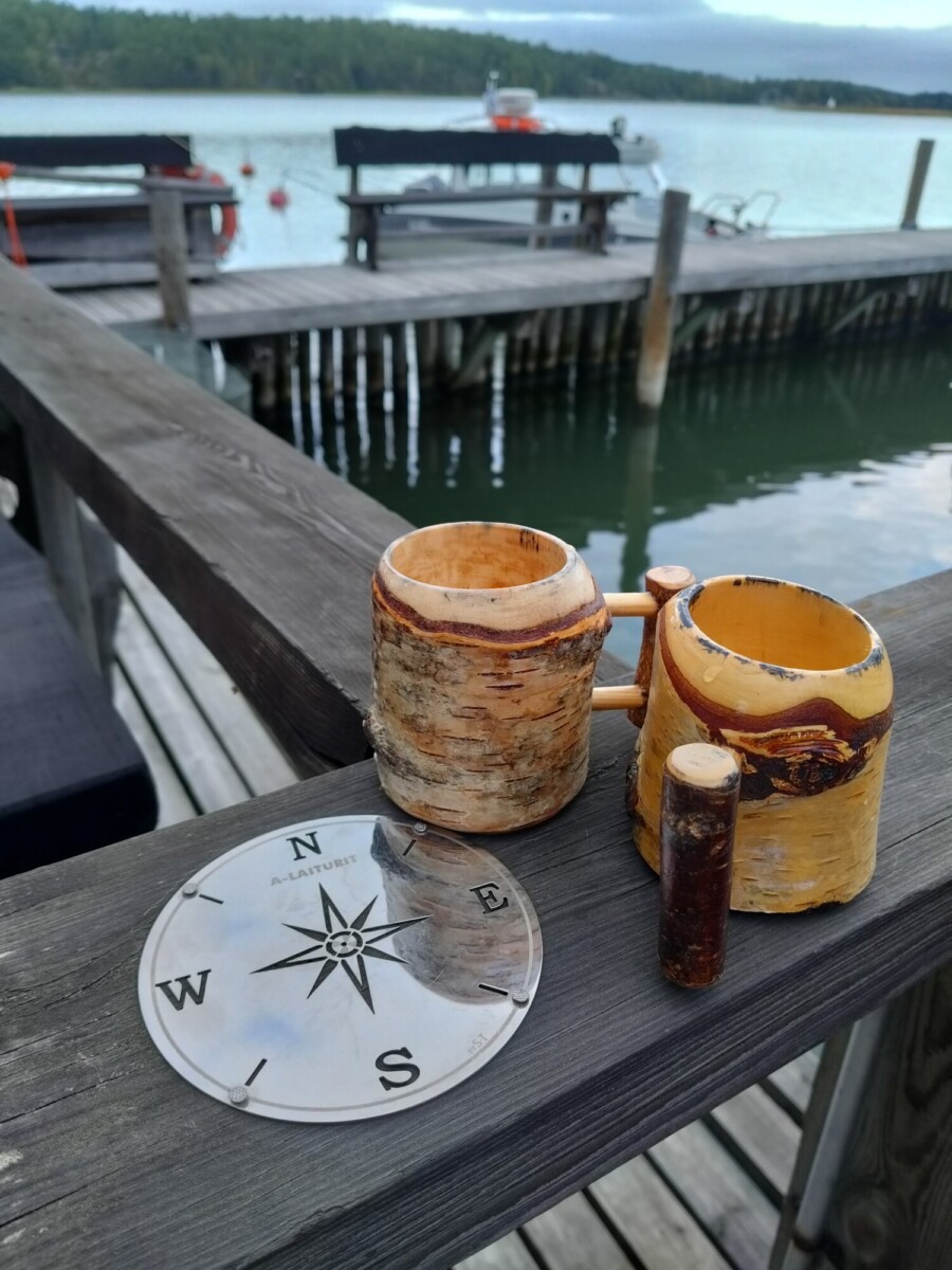 Kaksi puista mukia, pieni puinen sylinteri ja metallinen kompassilautanen on sijoitettu puukaiteelle järven rannalle, jonka taustalla näkyy laitureita, penkkejä, veneitä ja puita pilvisen taivaan alla.